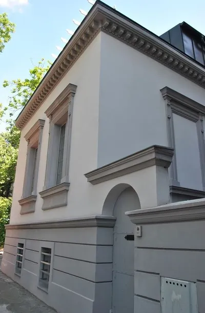 Bela kuća u klasičnom stilu sa ukrasnim vencem ispod krova, okvirima oko prozora i sivim podnožjem sa lajsnama