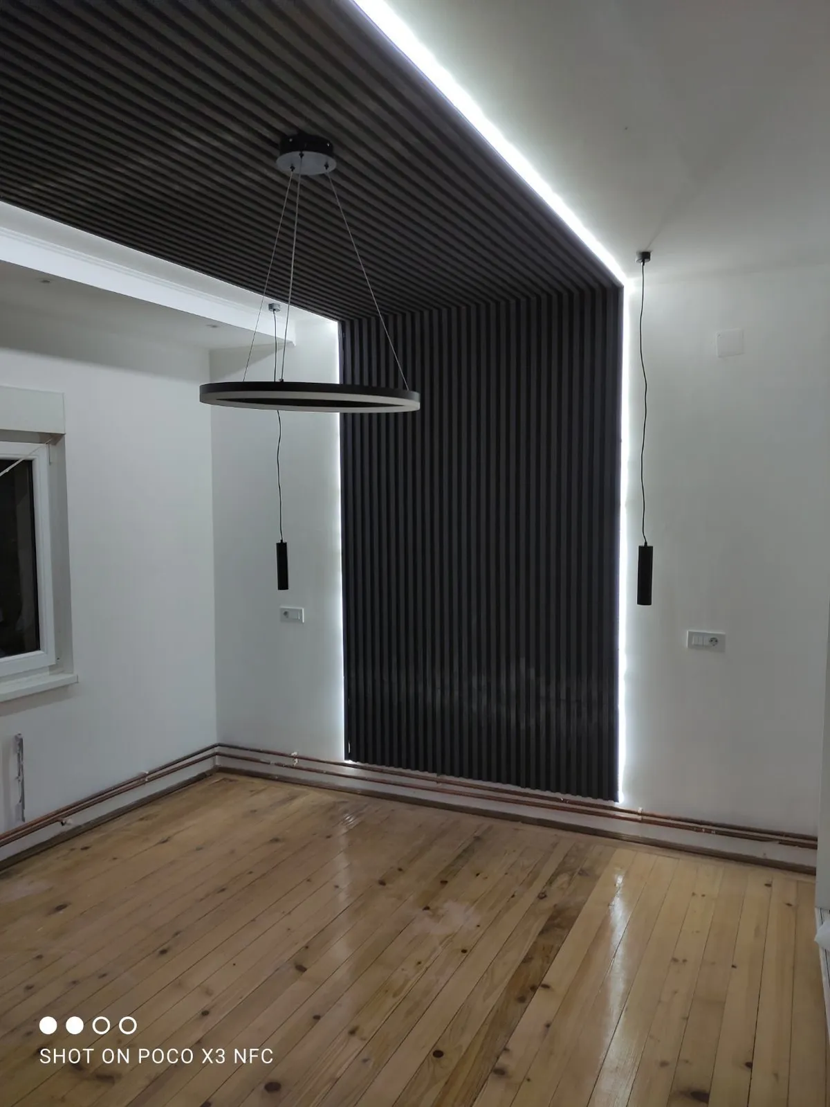 Crne letvice od poda preko plafona i viseće lampe Soba sa crnim letvicama od poda preko plafona, LED trakom koja ih uokviruje, visećim lampama i lusterom prstenom