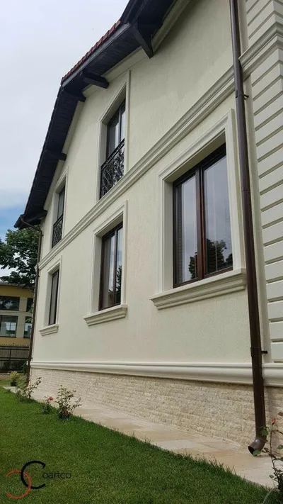 Kuća na sprat sa svetlom fasadom, okvirima oko prozora, vencem između spratova i kamenim podnožjem