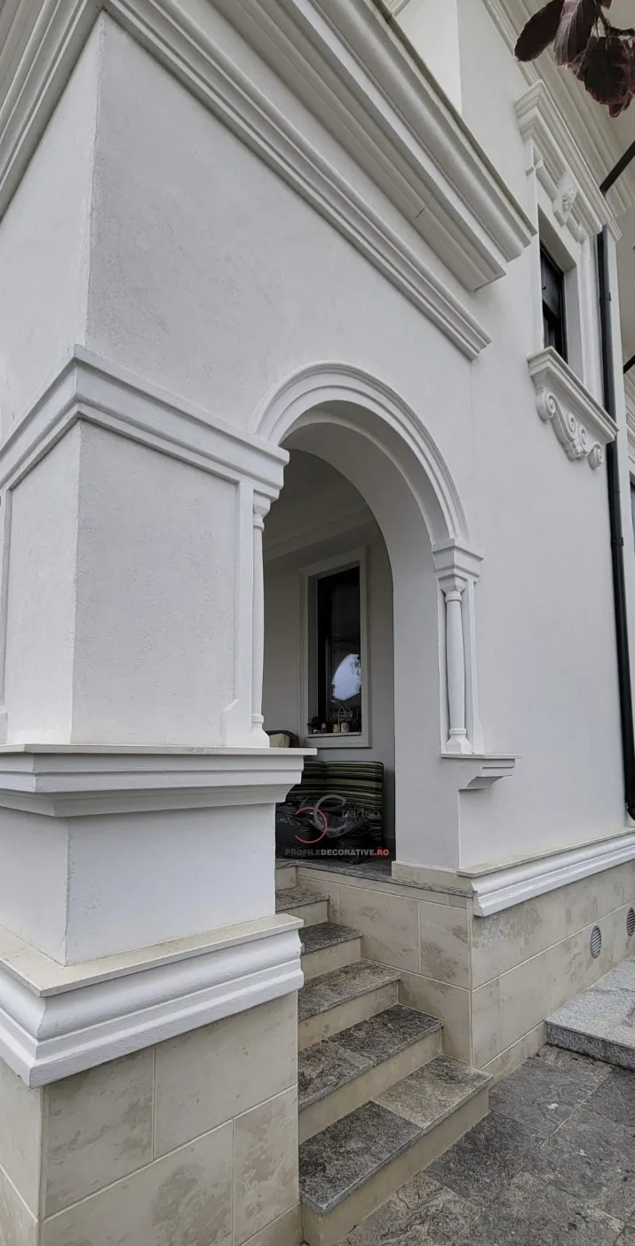 Lučni ulaz kuće sa pilastrima i fasadnim lajsnama Bela fasada sa lučnim ulazom uokvirenim lajsnama, pilastrima sa strane i vencem iznad, stepenice od kamena