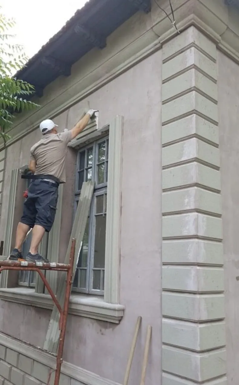 Postavljanje fasadne lajsne oko prozora na skeli Majstor na skeli postavlja fasadnu lajsnu oko prozora, a pored su već postavljene kocke na uglu kuće