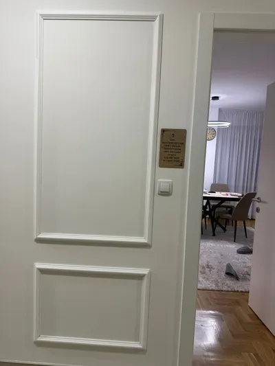 Beli zid sa ram lajsnama pored otvorenih vrata, pogled u dnevnu sobu sa trpezarijom