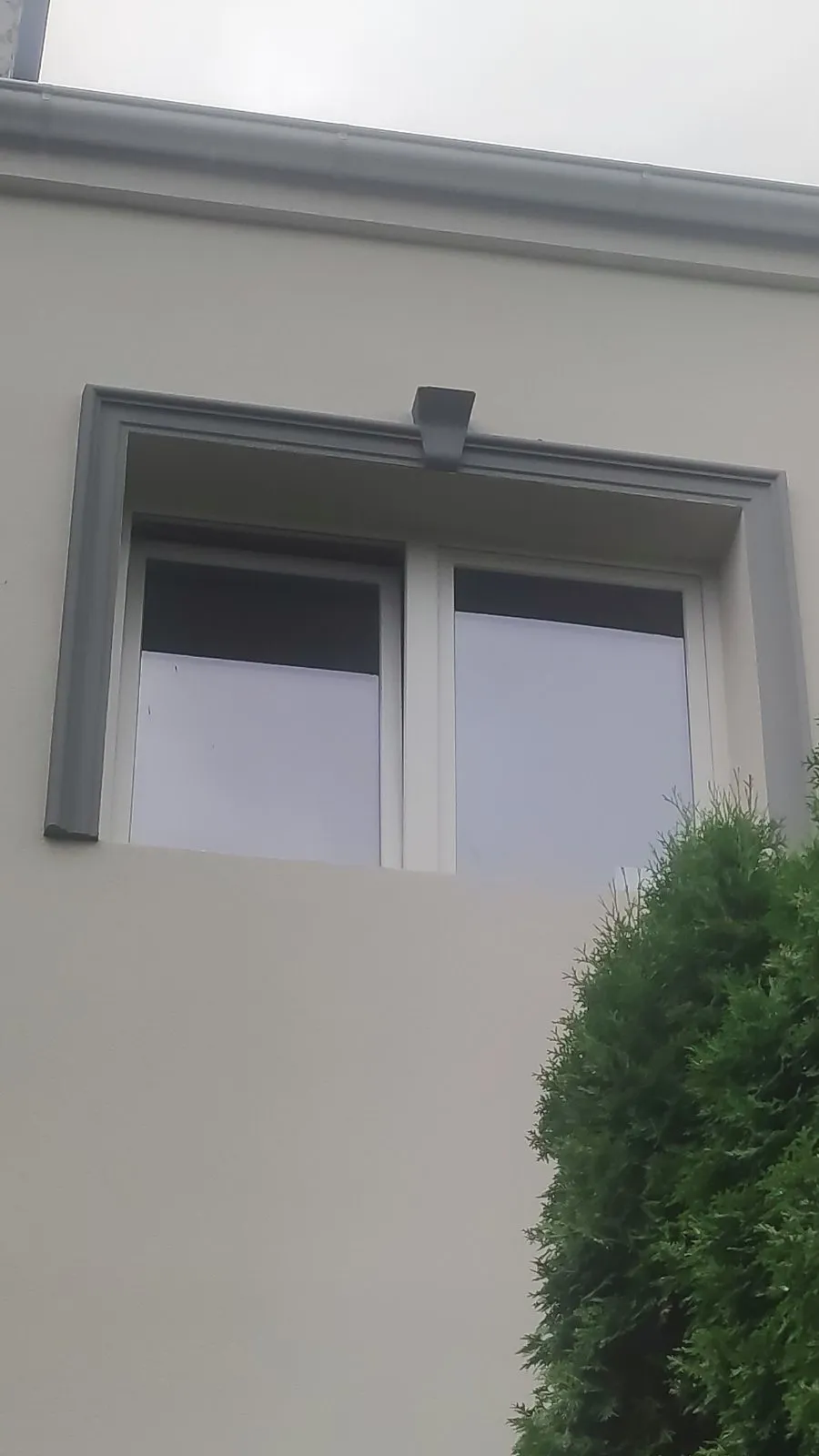 Sivi okvir prozora sa ključem na svetloj fasadi Prozor sa sivim okvirom od stiropora i ukrasnim ključem na vrhu, na svetloj fasadi iza tuje